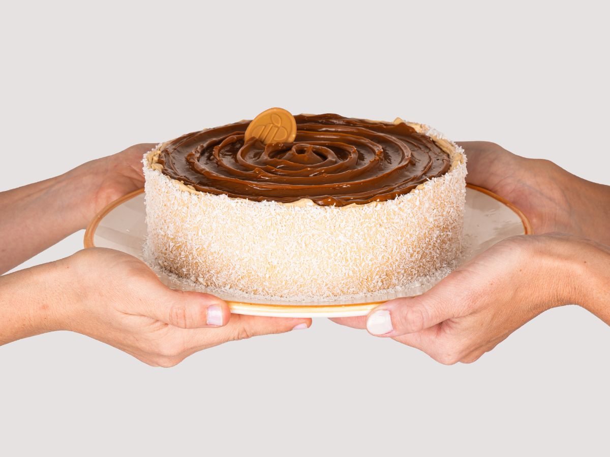 Bolo de coco com ameixa e doce de leite, segurado entre as mãos, com placa da Mombee